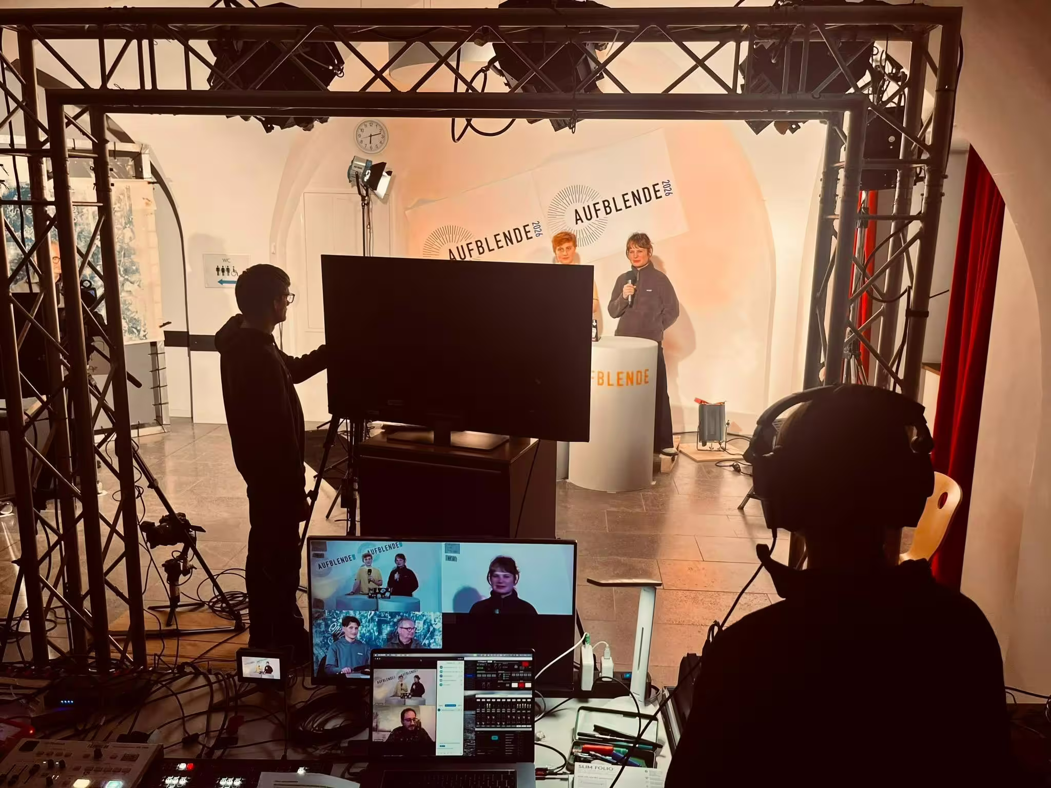 AUFBLENDE 2026 Live-Setup mit Monitoren, Technikcrew und Moderator*innen auf Bühne hinter Truss-Rahmen bei Veranstaltung