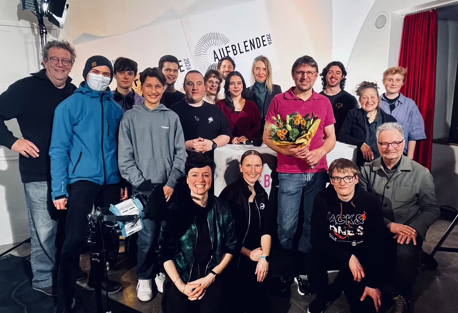 Gruppenfoto des AUFBLENDE2026 Teams und Teilnehmenden vor Banner, lächelnd und Blumenstrauß überreicht bei Event