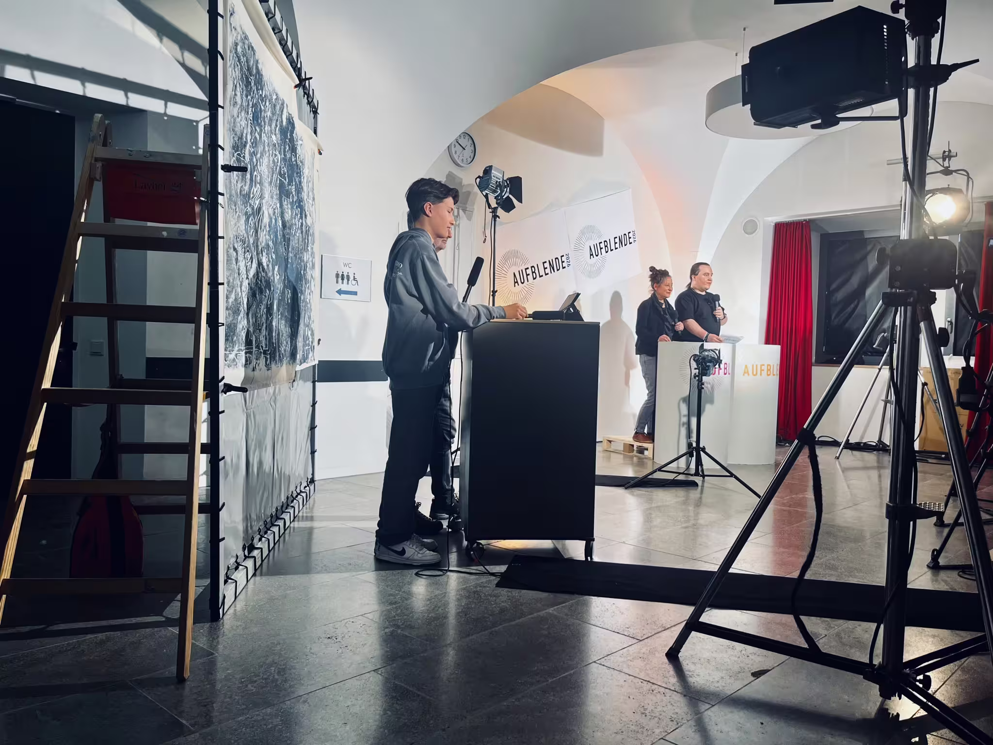 Teenager spricht am Pult bei AUFBLENDE 2026 Veranstaltung, Bühnenlicht, Mikrofone und Kameraausrüstung im Raum