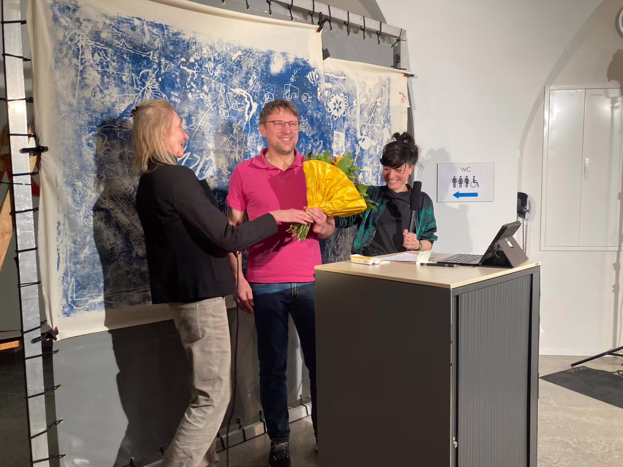 Drei lächelnde Menschen bei AUFBLENDE2026: Mann erhält Blumen vor blau-weißem Kunstwerk bei Preisverleihung (Eventfoto)