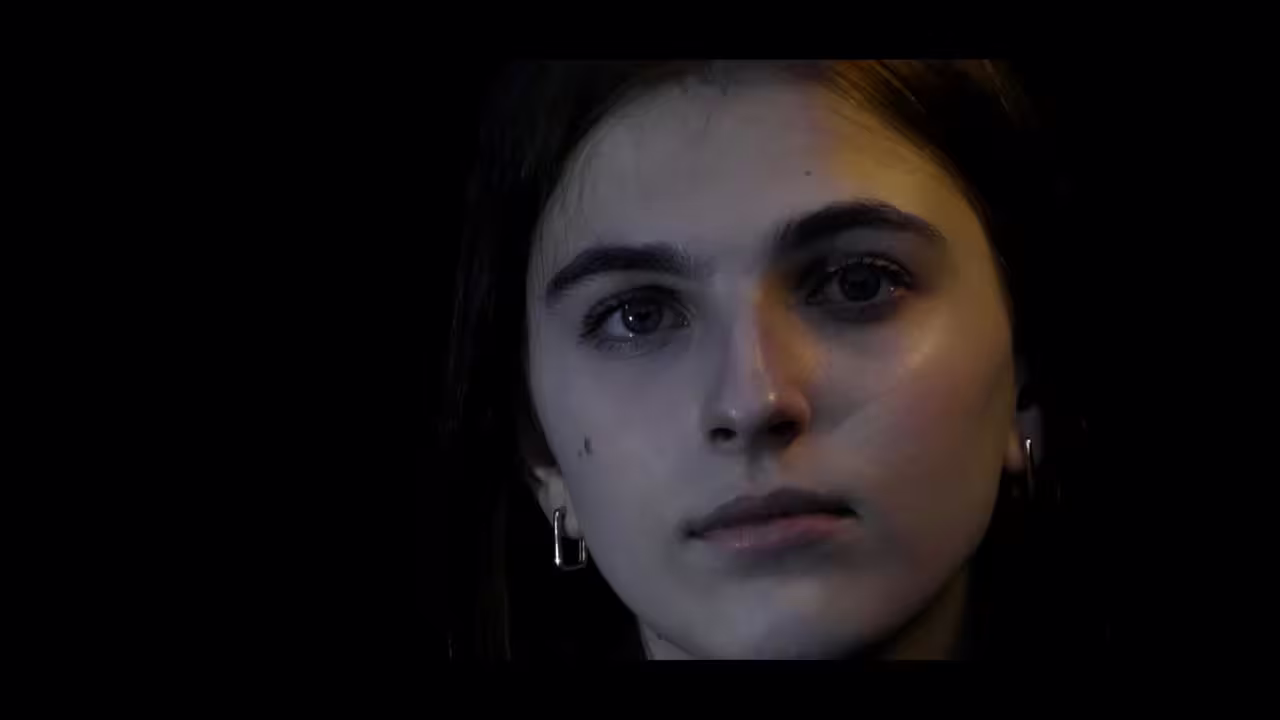 Nahaufnahme eines jungen Mädchens mit ernster Miene vor dunklem Hintergrund, Filmstill aus BRUCH (AUFBLENDE2026)